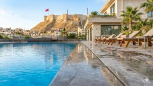 Gündüz çekilmiş, berrak suyuyla parlayan modern bir taşmalı havuzun kenardan görünümü. Sağ tarafta lüks bir villa ve güneşlenme şezlongları, arka planda ise tarihi Gaziantep Kalesi yükseliyor.